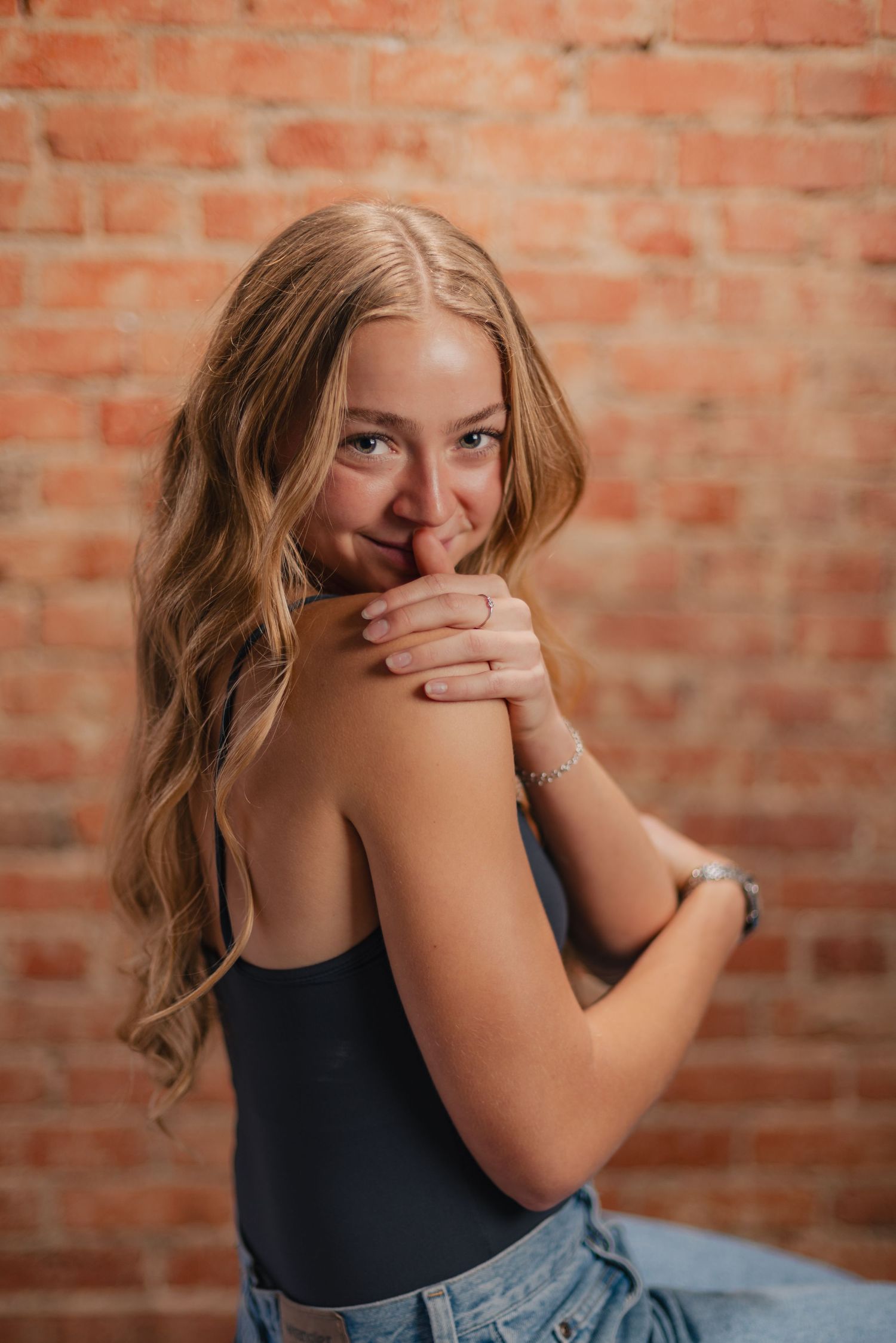 Person in dark tank top and jeans poses playfully against brick wall background.