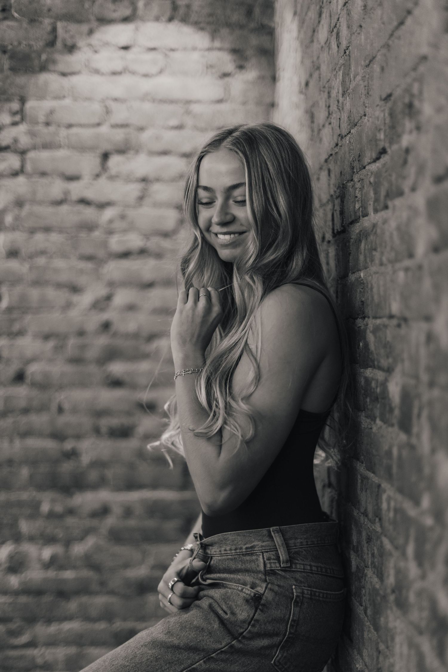 Black and white portrait of a person smiling against a brick wall wearing casual attire.