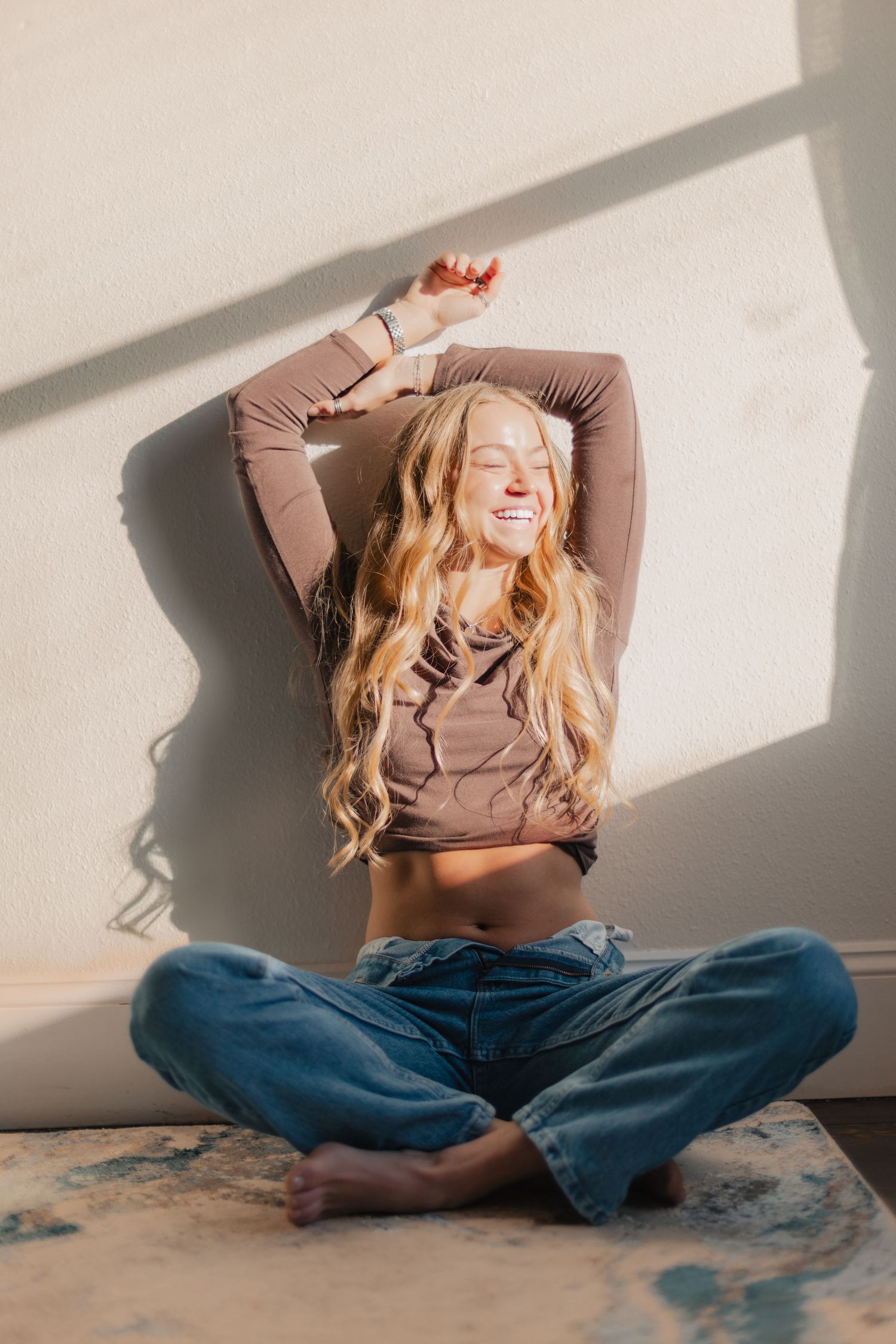 Candid lifestyle portrait of a person in a brown sweater and jeans sitting cross-legged against a white wall in sunlight.