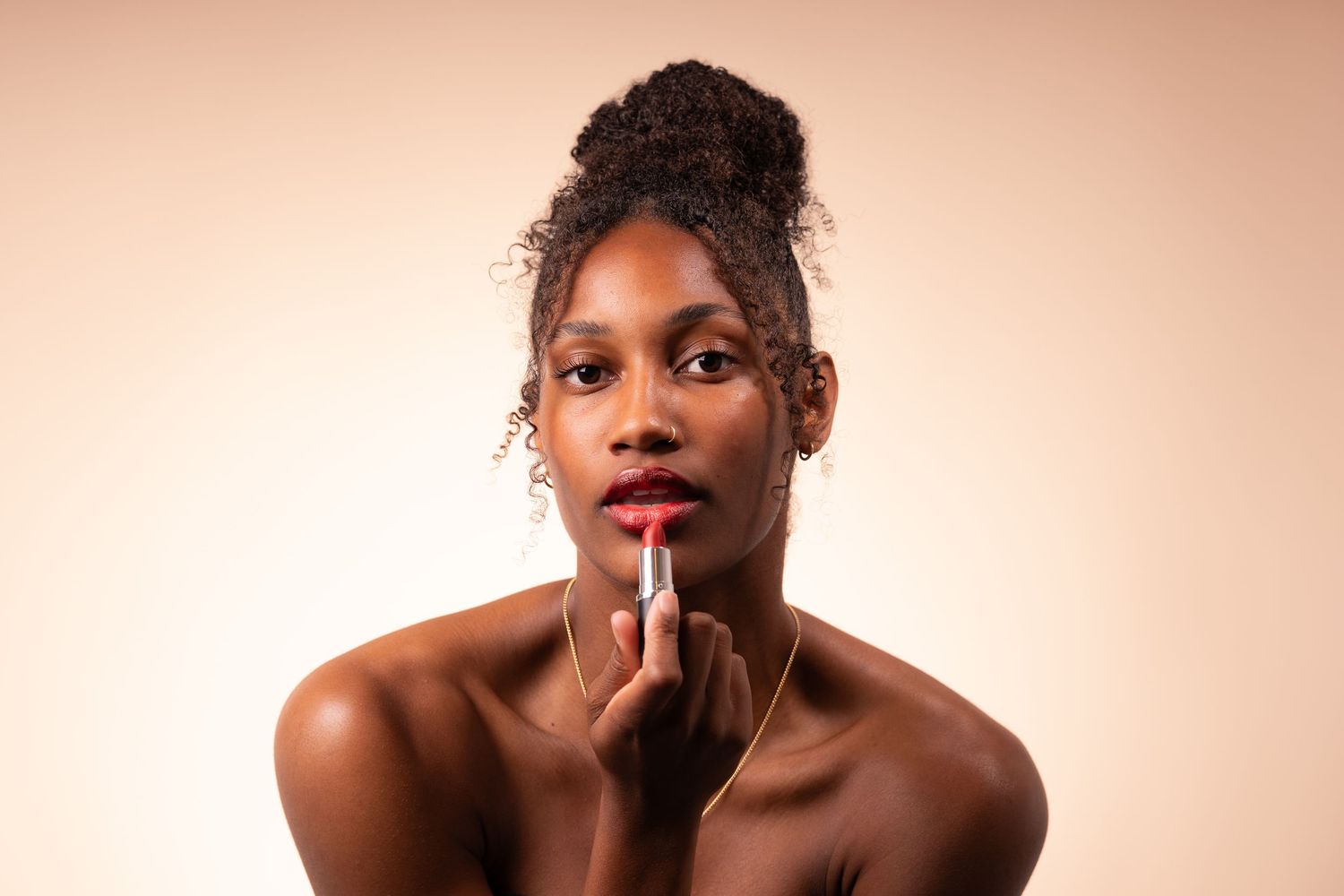 Beauty portrait of a person applying bright red lipstick against a warm peach background.