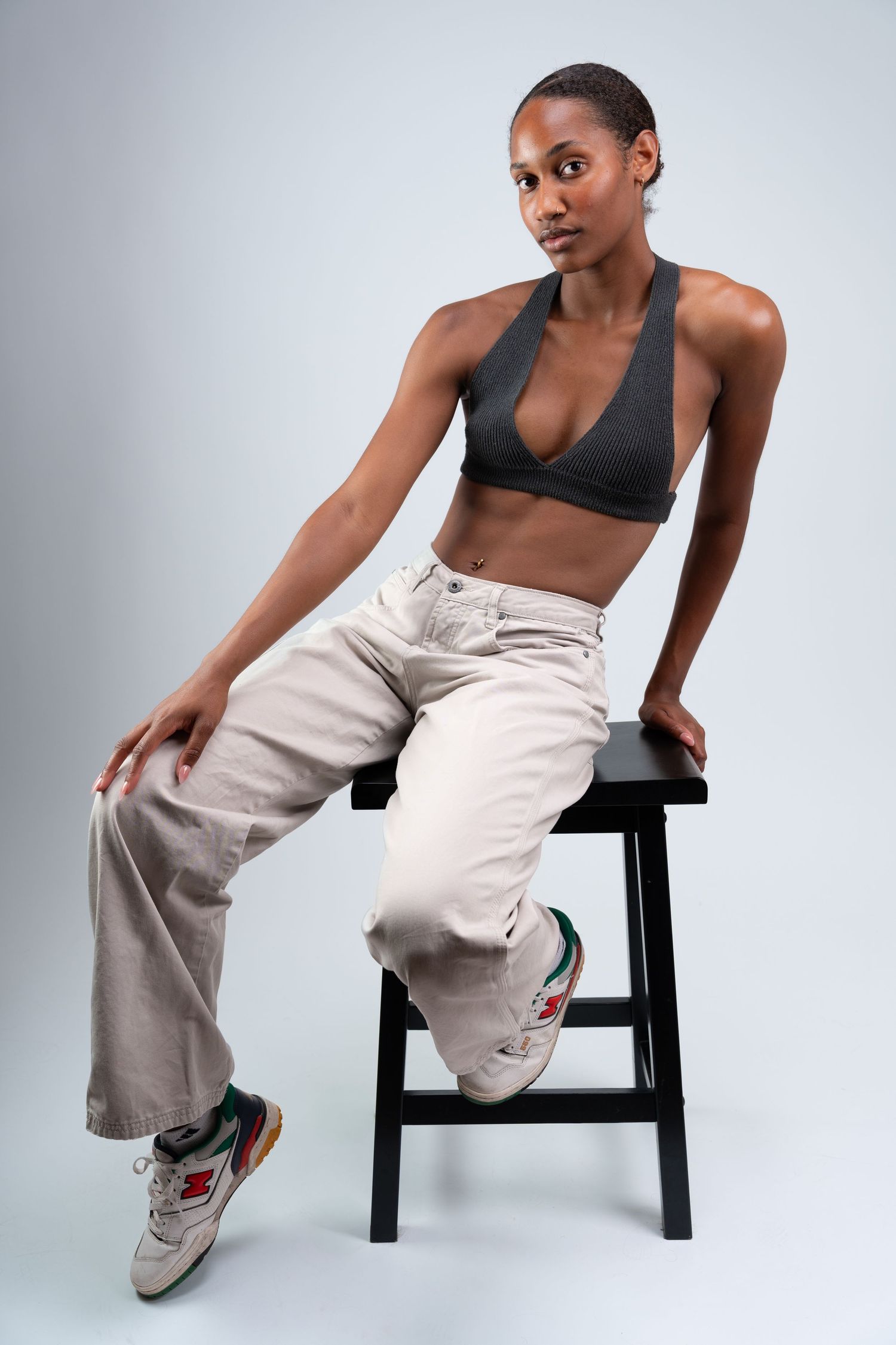 Athletic figure in black sports bra and beige pants strikes dynamic poses while seated on black stool against gray backdrop.