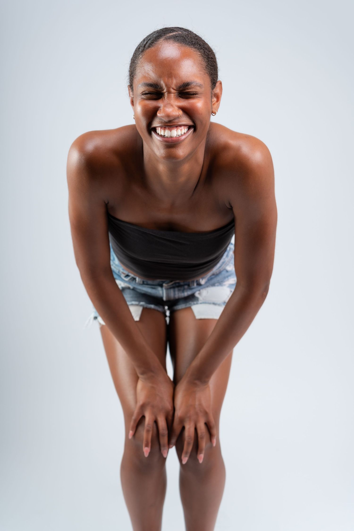 A joyful person wearing a black top and denim shorts bends forward with hands on knees against a white background.