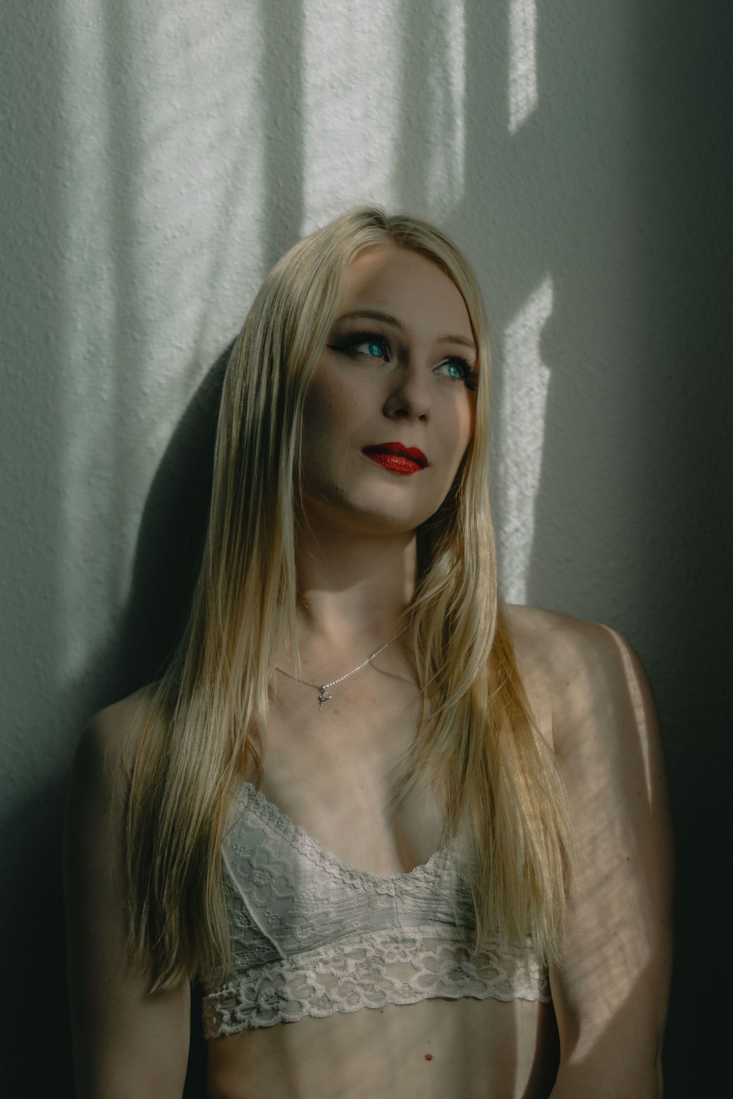 Blonde woman with red lips and blue eyes wearing lace bralette and necklace, leaning against wall in moody light.
