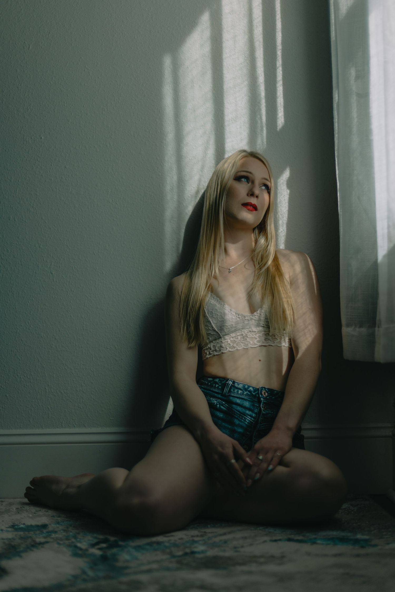 Blonde woman in lace bralette and teal shorts sitting on floor against wall in dramatic window light.