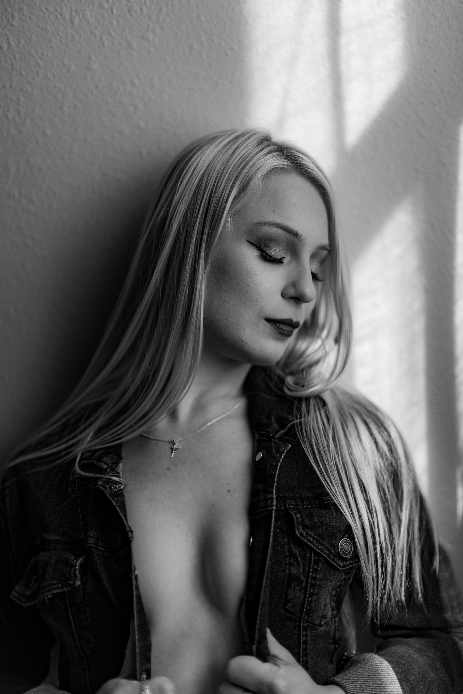 Black and white portrait of a blonde woman in a denim jacket leaning against a wall with soft window light.