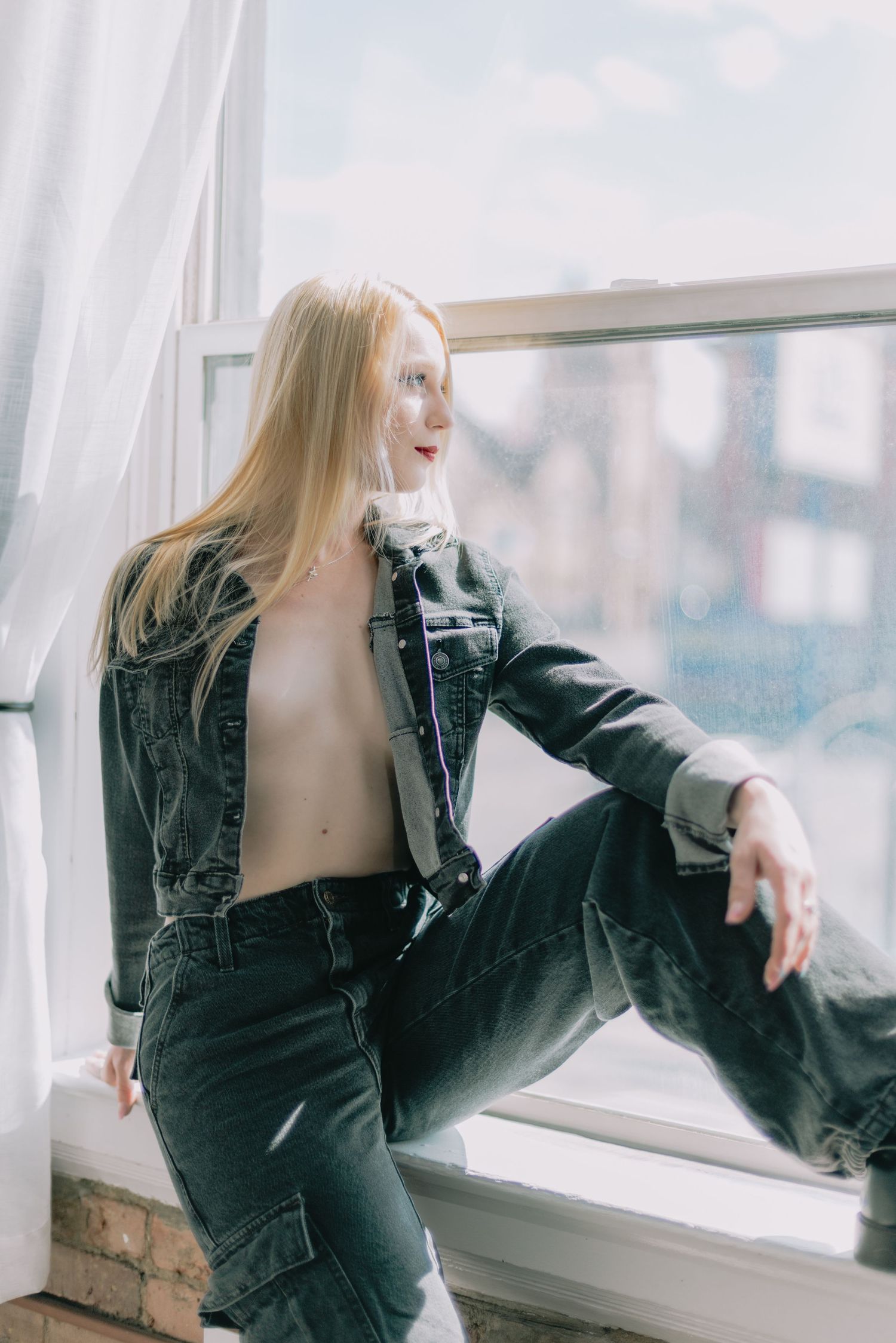 Young blonde woman in denim jacket and jeans posing by a large window with sheer curtains.