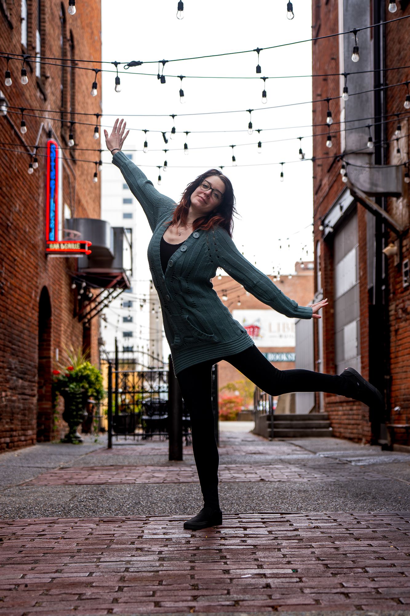 A dancer performs graceful poses on a cobblestone street between brick buildings with string lights overhead.