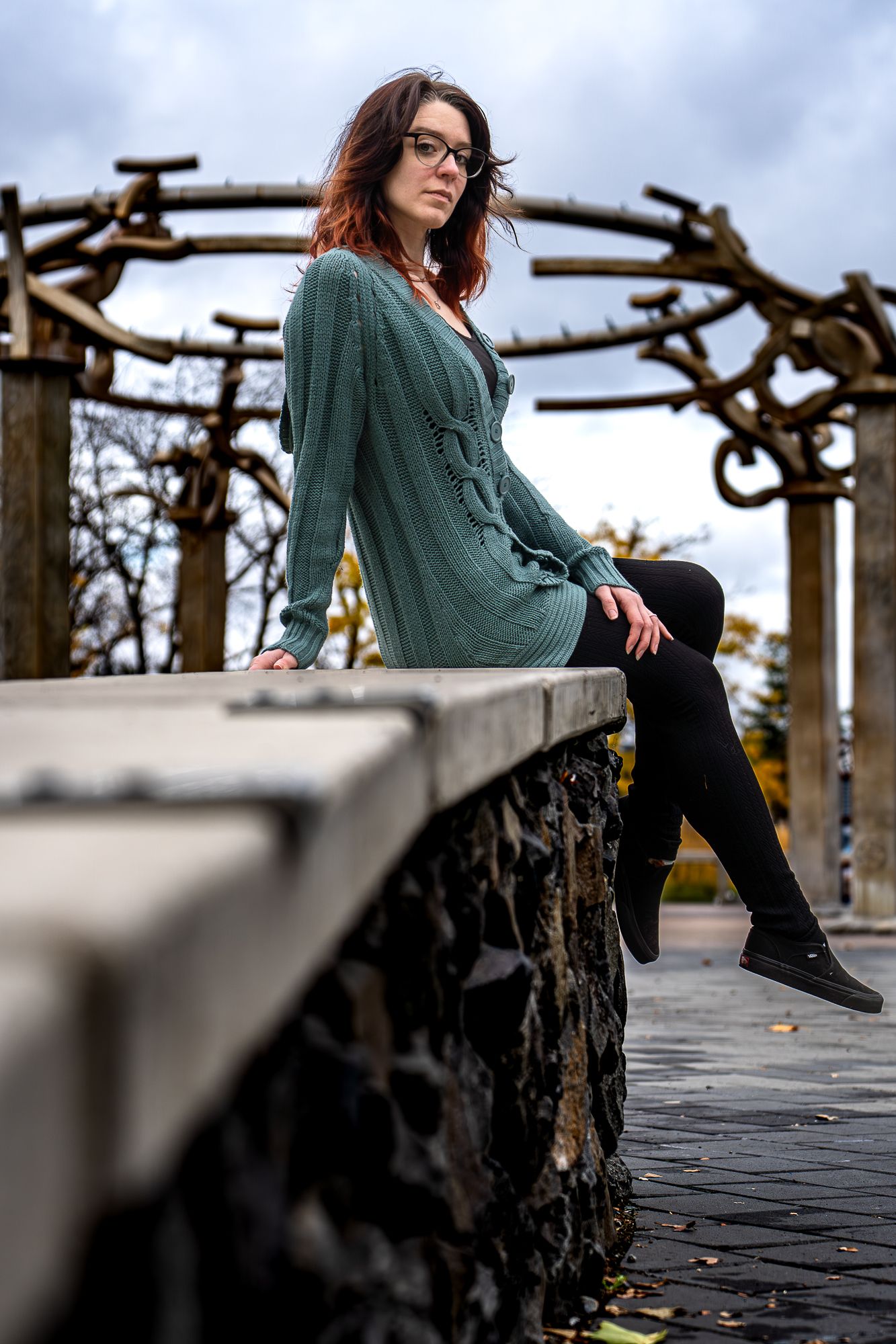 Person in a green knit sweater and glasses sits on a stone ledge near an ornate metal arbor structure.