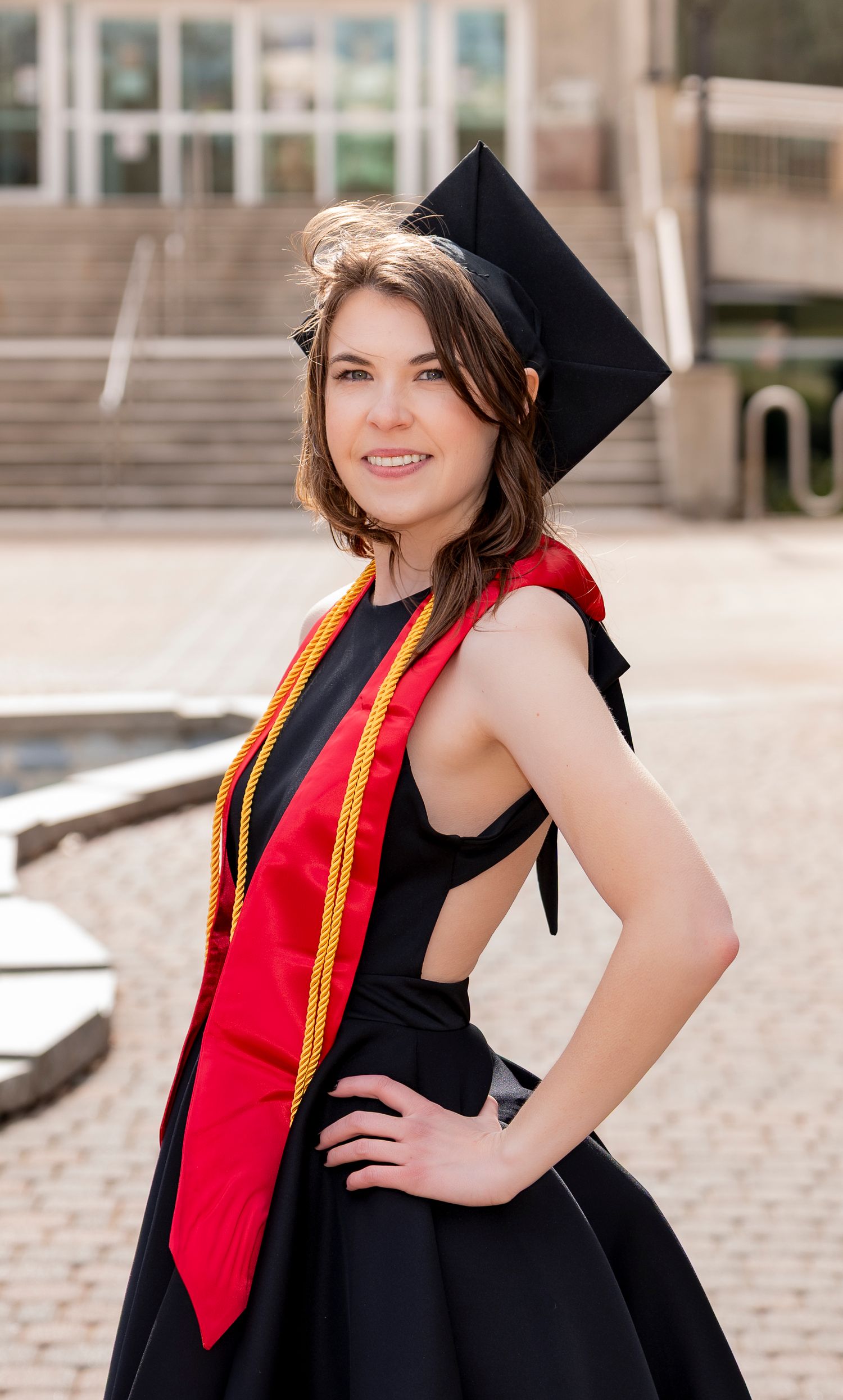 A graduate in a black cap and gown with red stole poses outdoors for a series of graduation portraits.