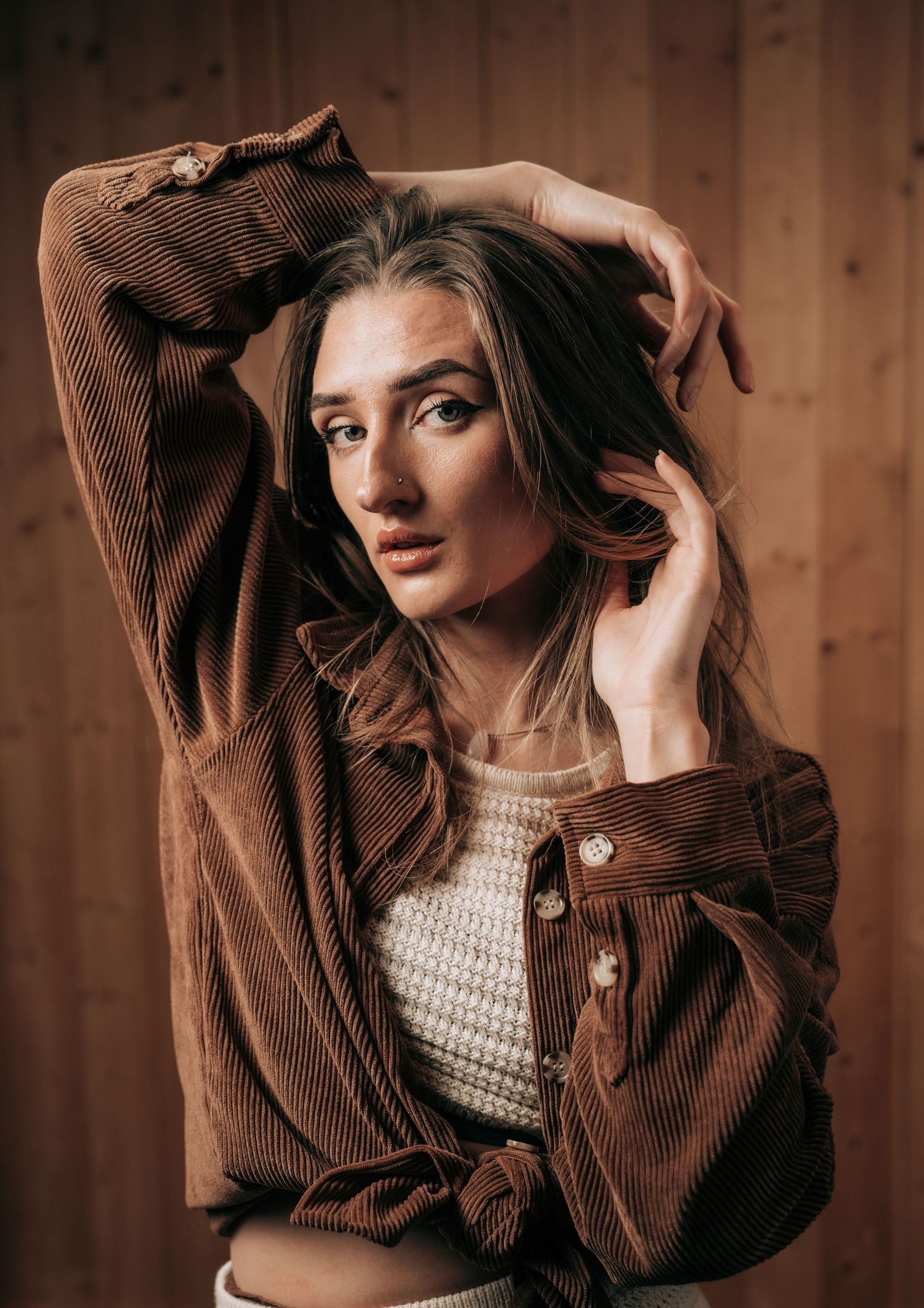 Moody portrait of a model wearing a brown corduroy jacket and cream sweater against wooden paneled background.