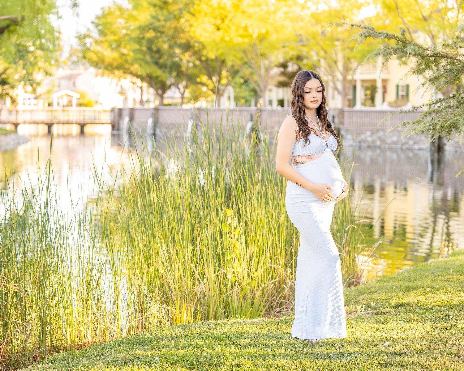 Maternity Photoshoot with a photographer using natural light. The expected mother poses beside a lake as light shines naturally on the water