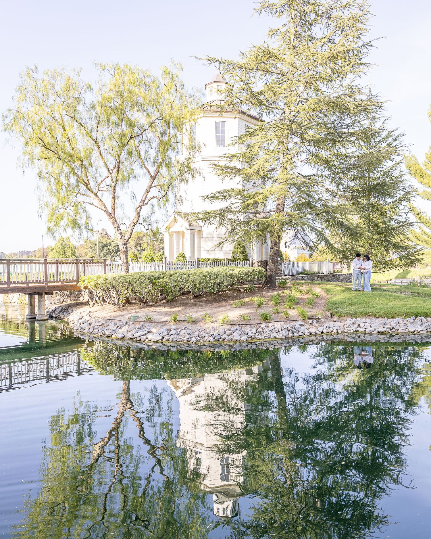 Couple poses for maternity photos. A lake allows for a reflection to appear in water of the couple. Photogapher uses natrual light for photo