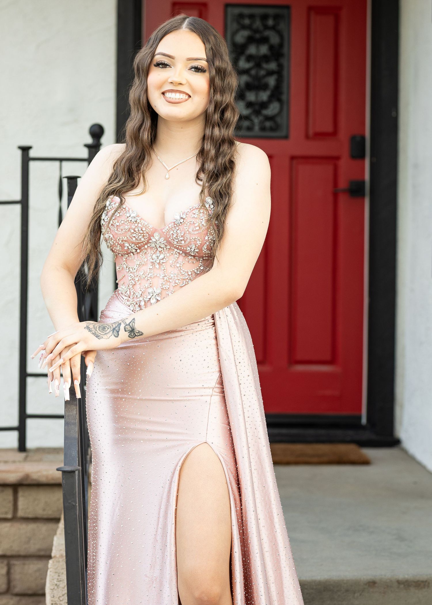 A female model in a blush pink formal gown with embellished bodice poses on steps by a red door.