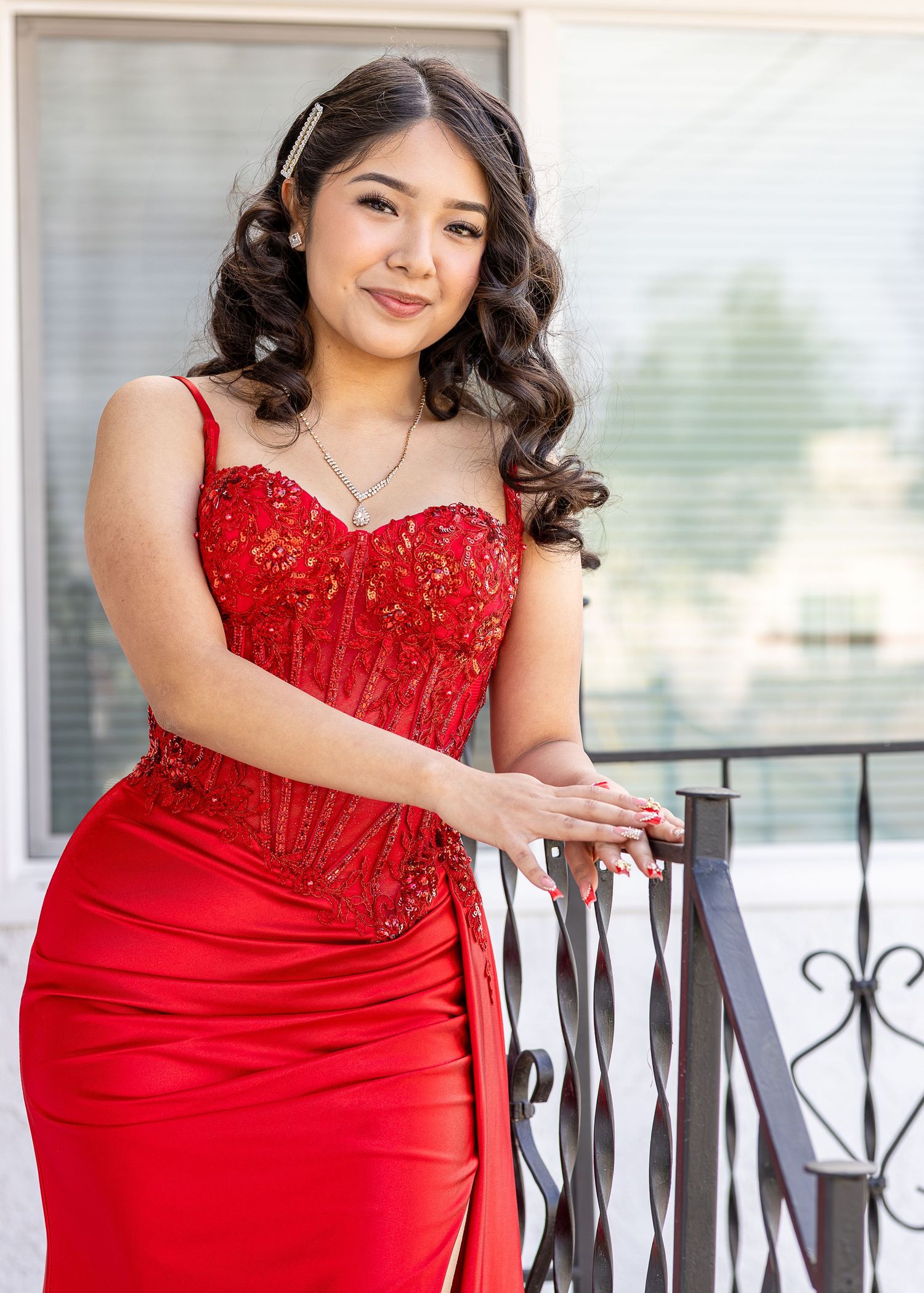 A woman models in a stunning red formal gown with a beaded bodice, poses elegantly by a metal railing in an outdoor setting for a portrait.