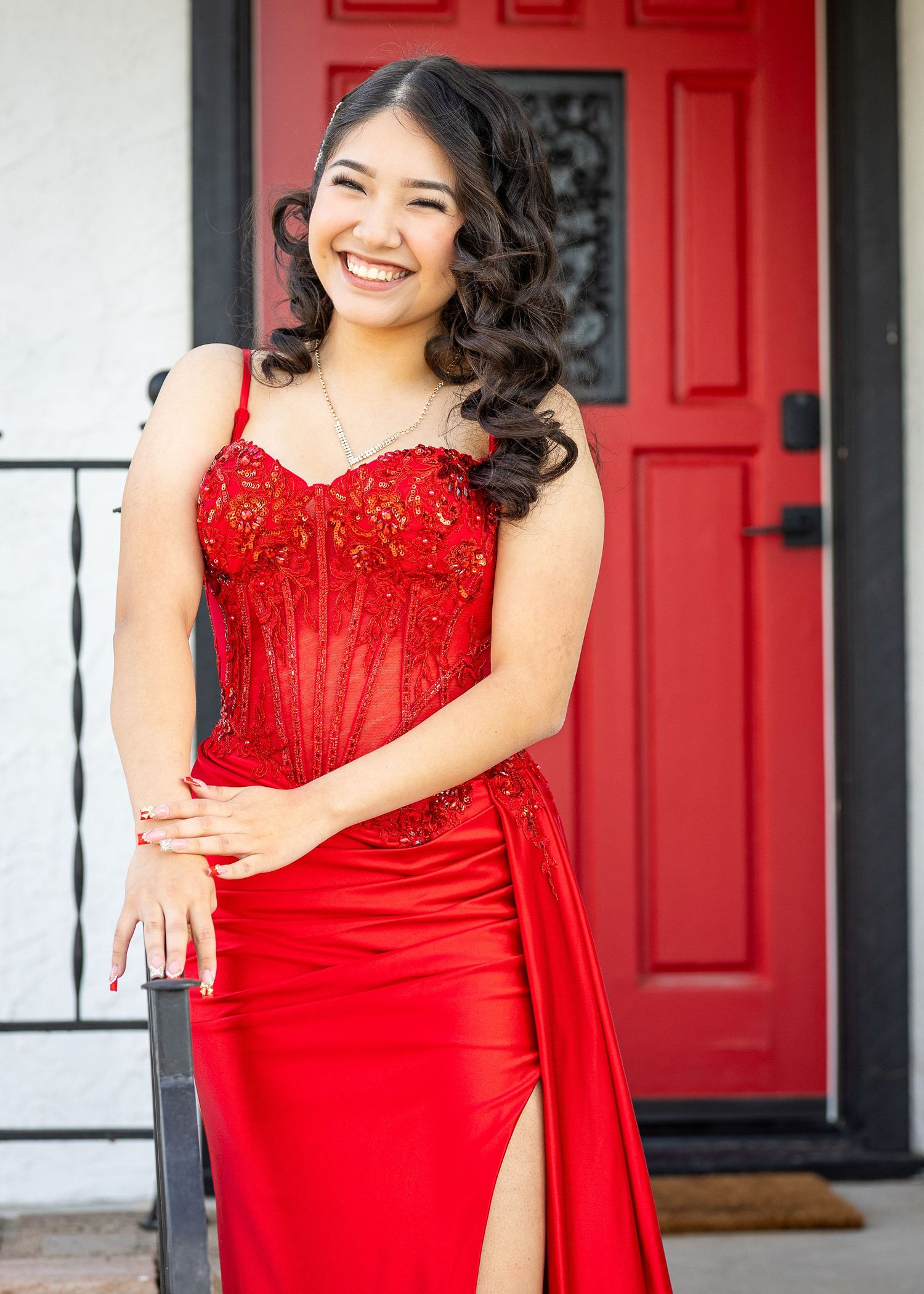 Elegant model in a glamorous red evening prom dress with beaded bodice poses cheerfully against a red door backdrop.