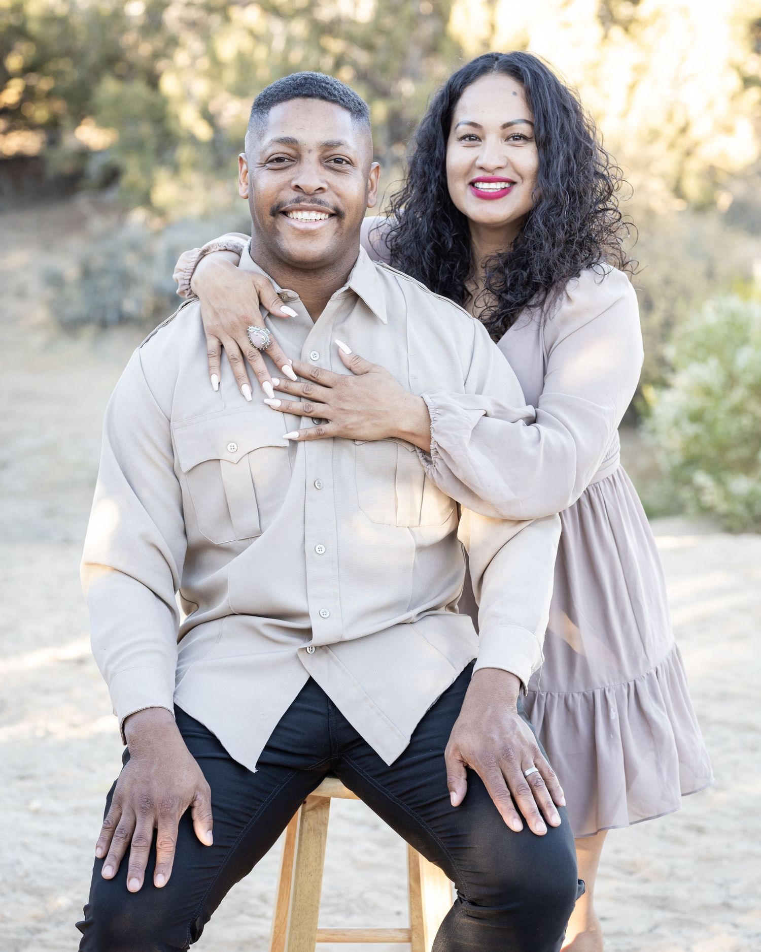 A portrait photo of a couple in beige outfits embracing while sitting on a wooden stool outdoors during golden hour lighting.