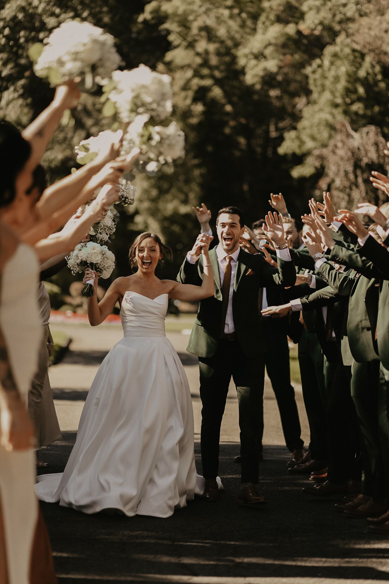 A wedding party celebrates outdoors with raised hands and white bouquets.
