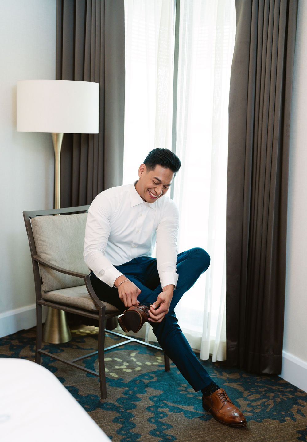 Well-dressed person in white shirt and blue pants putting on brown dress shoes in hotel room.