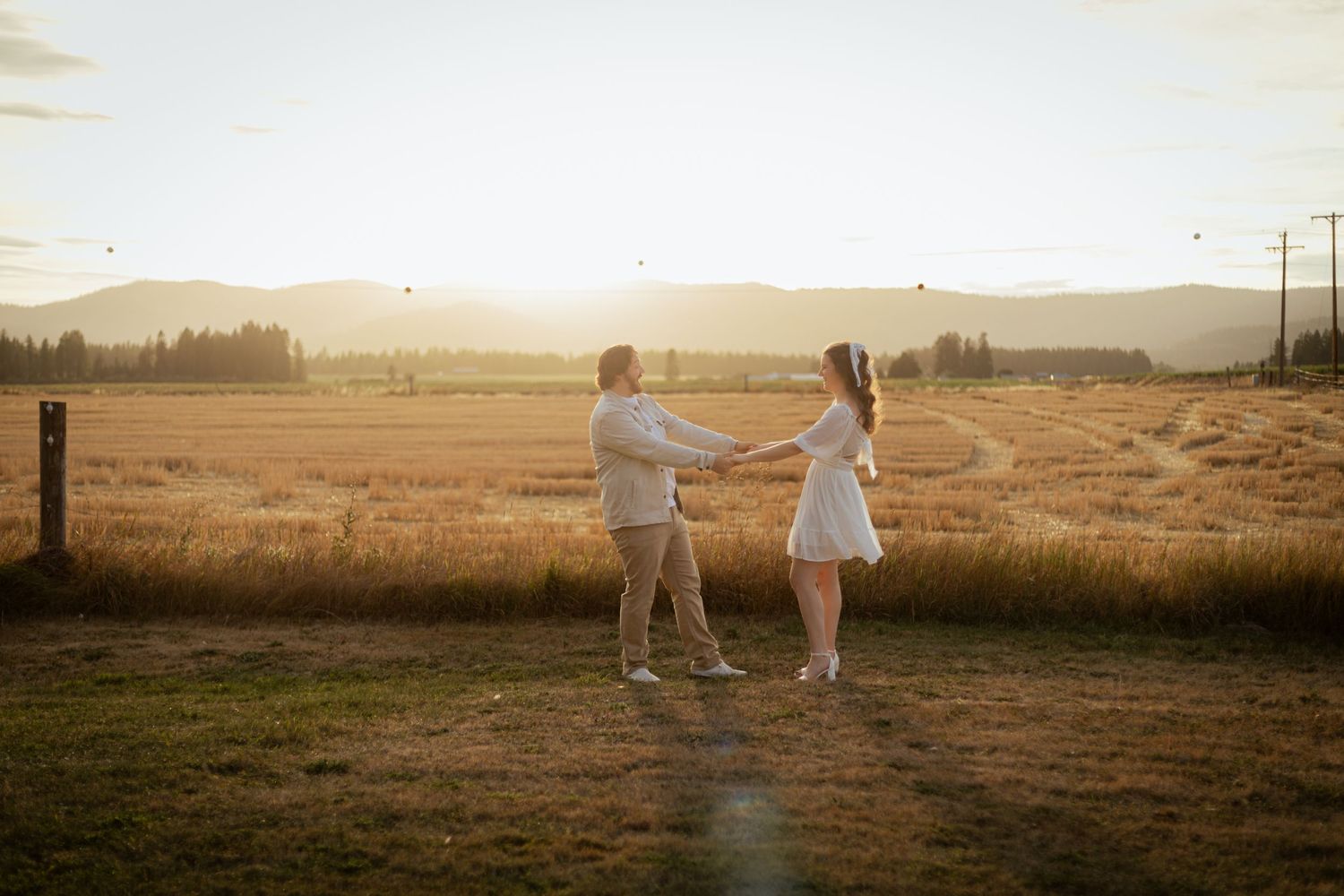A couple dances together in a golden field at sunset with mountains in the background.
