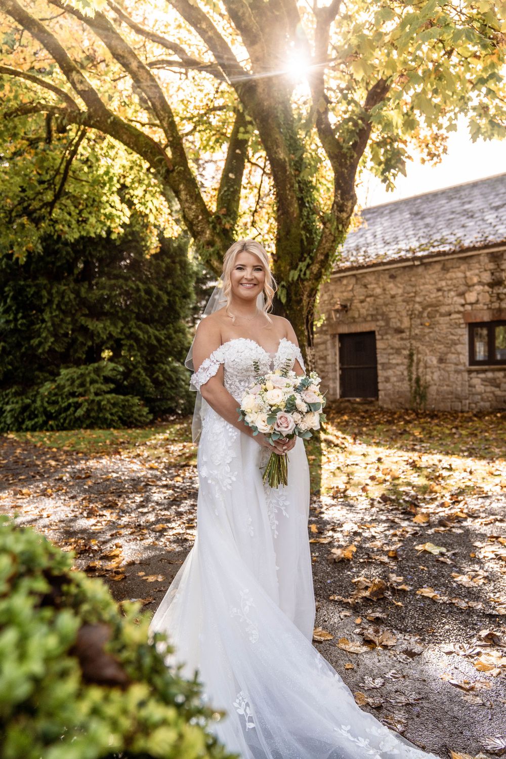Sunlit autumn wedding portrait with stone building and tree backdrop.
