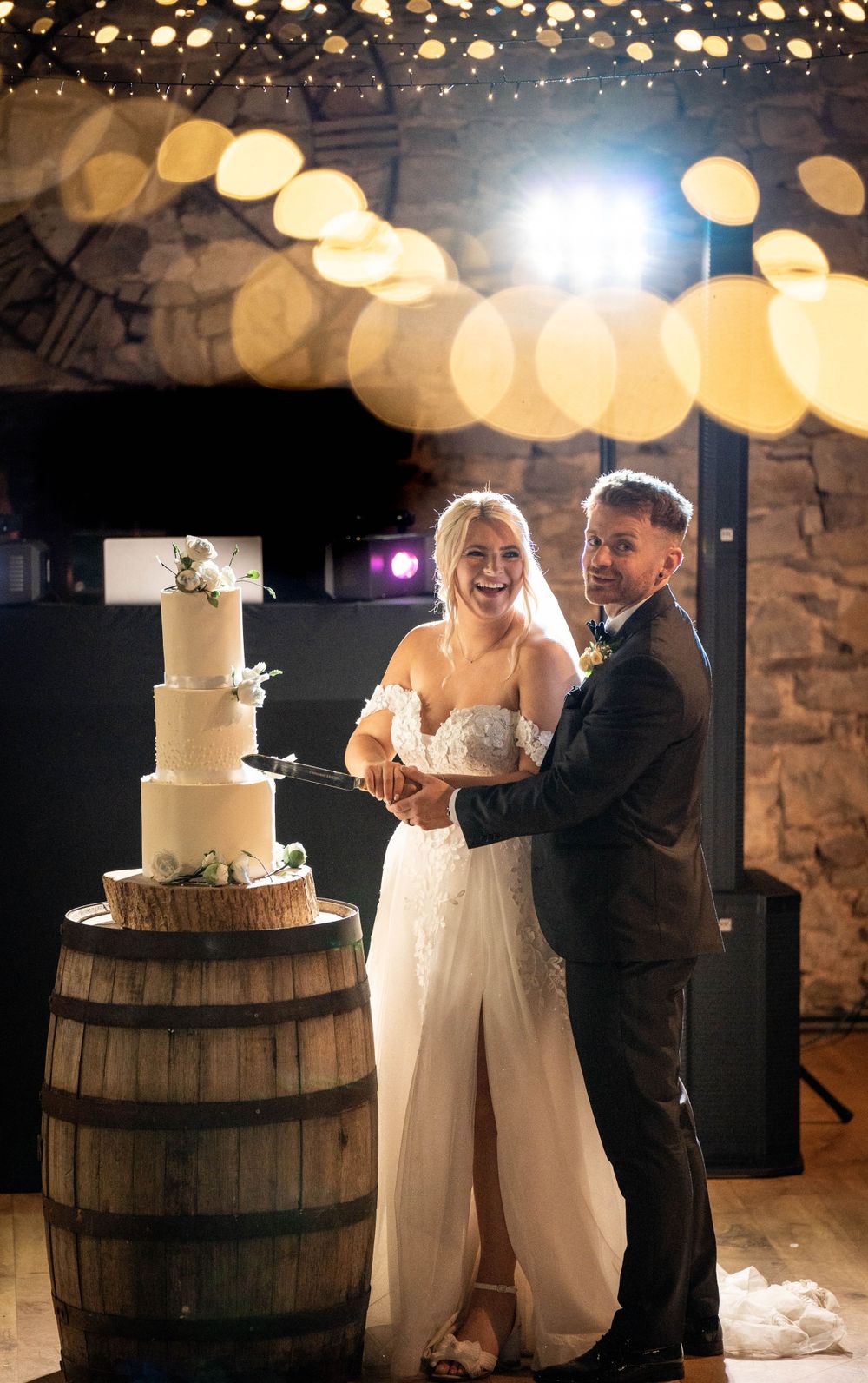 Wedding cake cutting moment beside rustic barrel under twinkle lights.