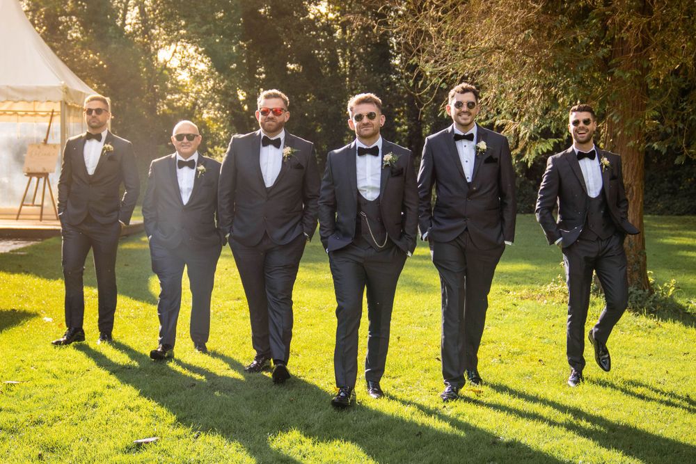 Groomsmen in black tuxedos walk confidently across a sunlit lawn with a wedding tent in the background.