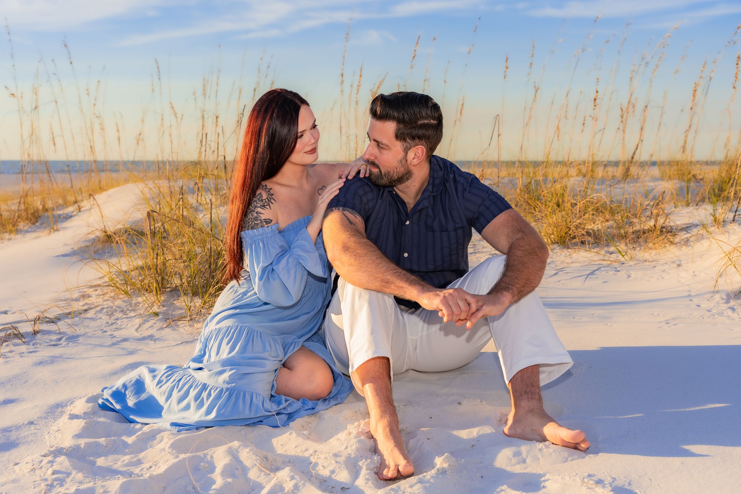 A couple sits together on white sand dunes at sunset with tall beach grass in the background.