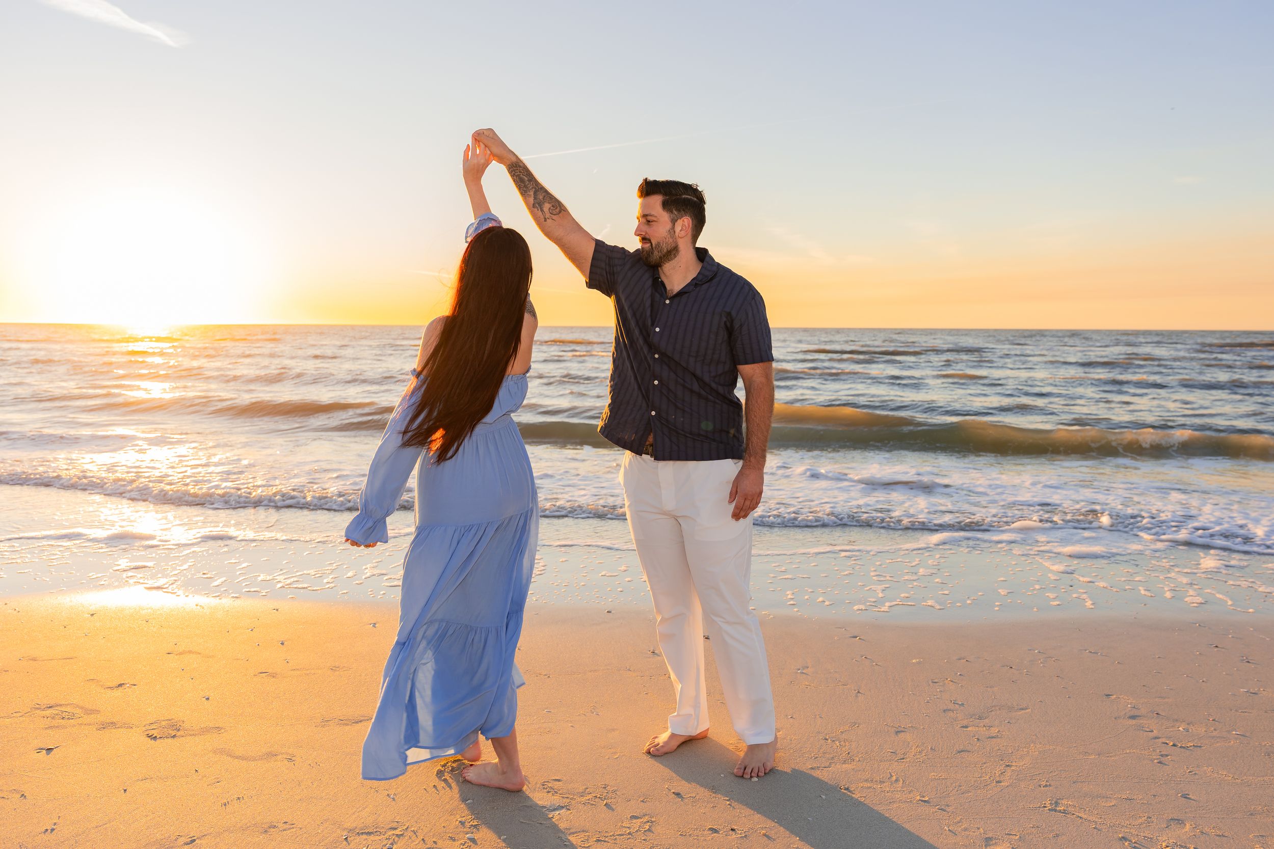 A couple dances together on a sandy beach at sunset with ocean waves in the background.