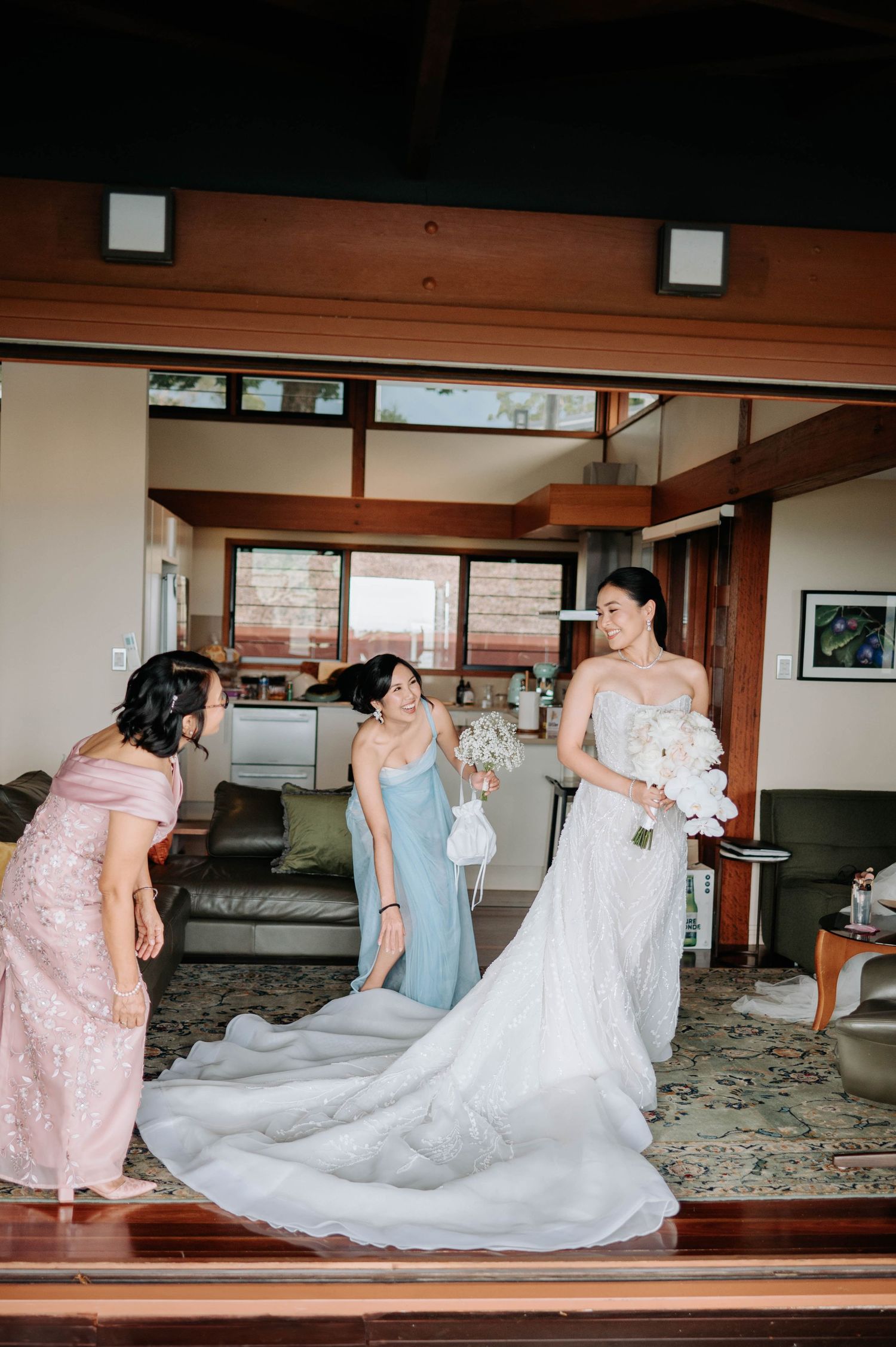 Bride in lace gown with long train gets final adjustments in luxurious interior setting.
