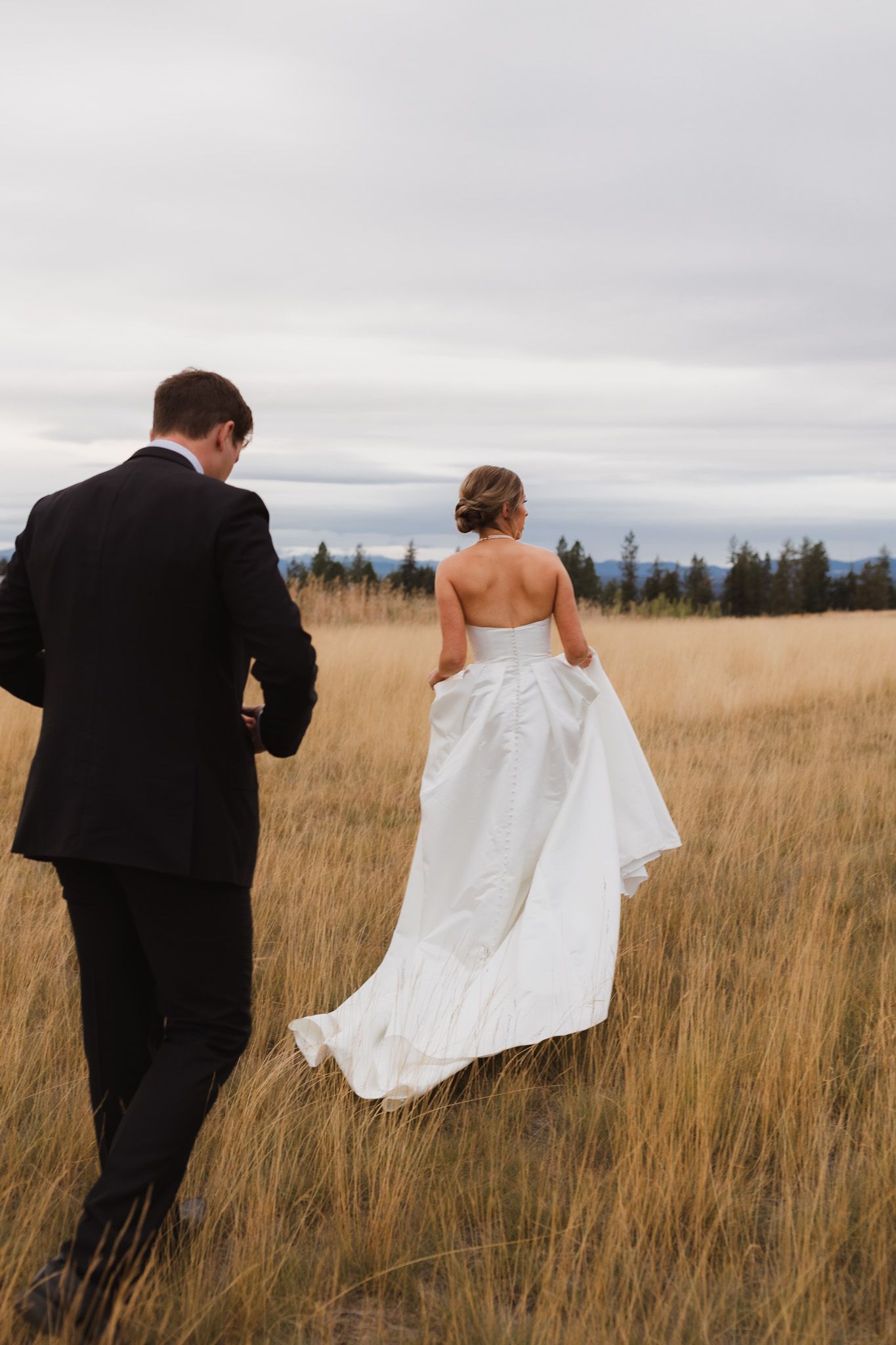 A couple walks through tall grass with mountains in the distance on their wedding day. Spokane wedding photographer.