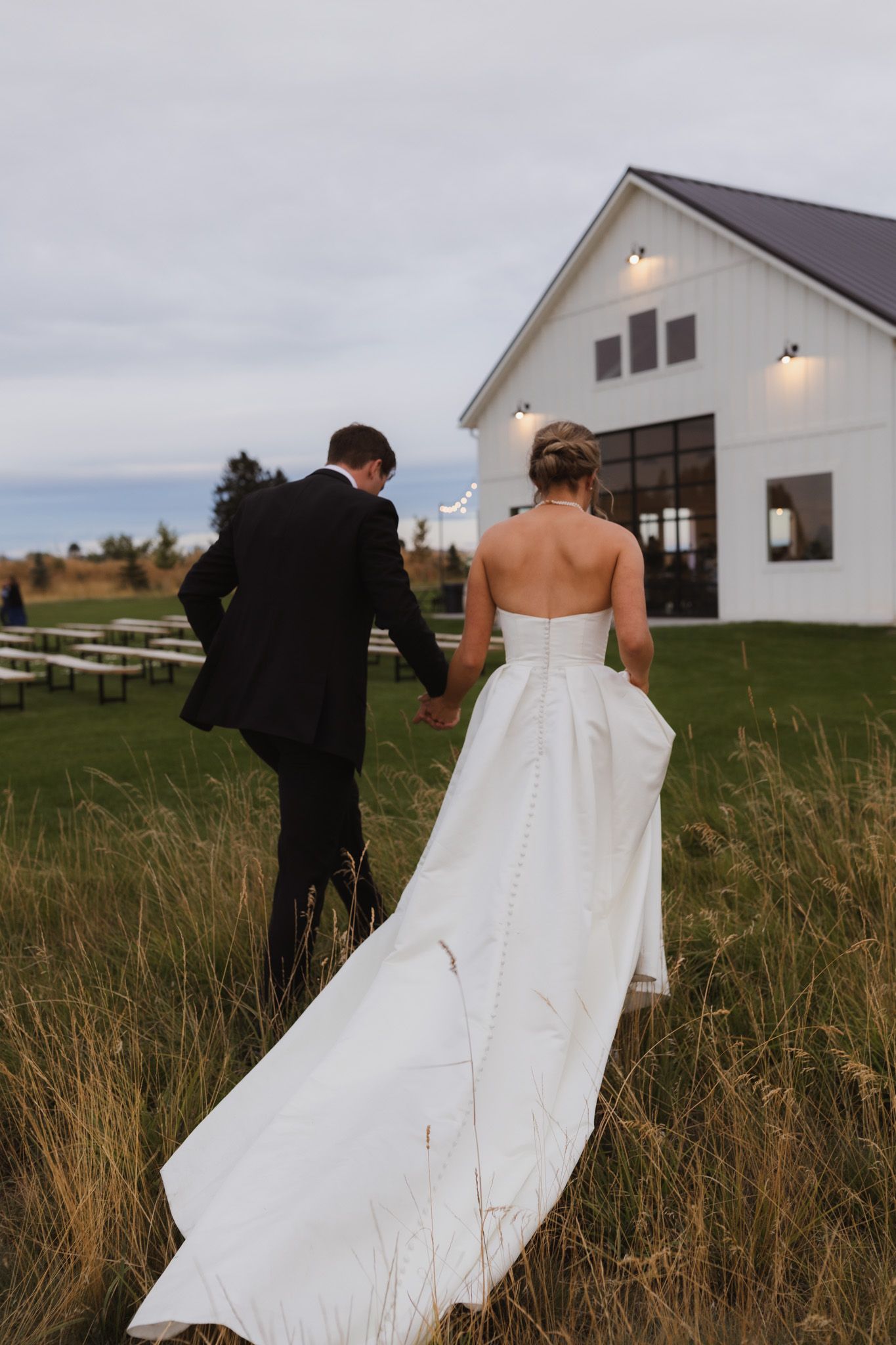 Newlyweds walking toward a modern white barn venue through tall grass. Spokane wedding photographer.