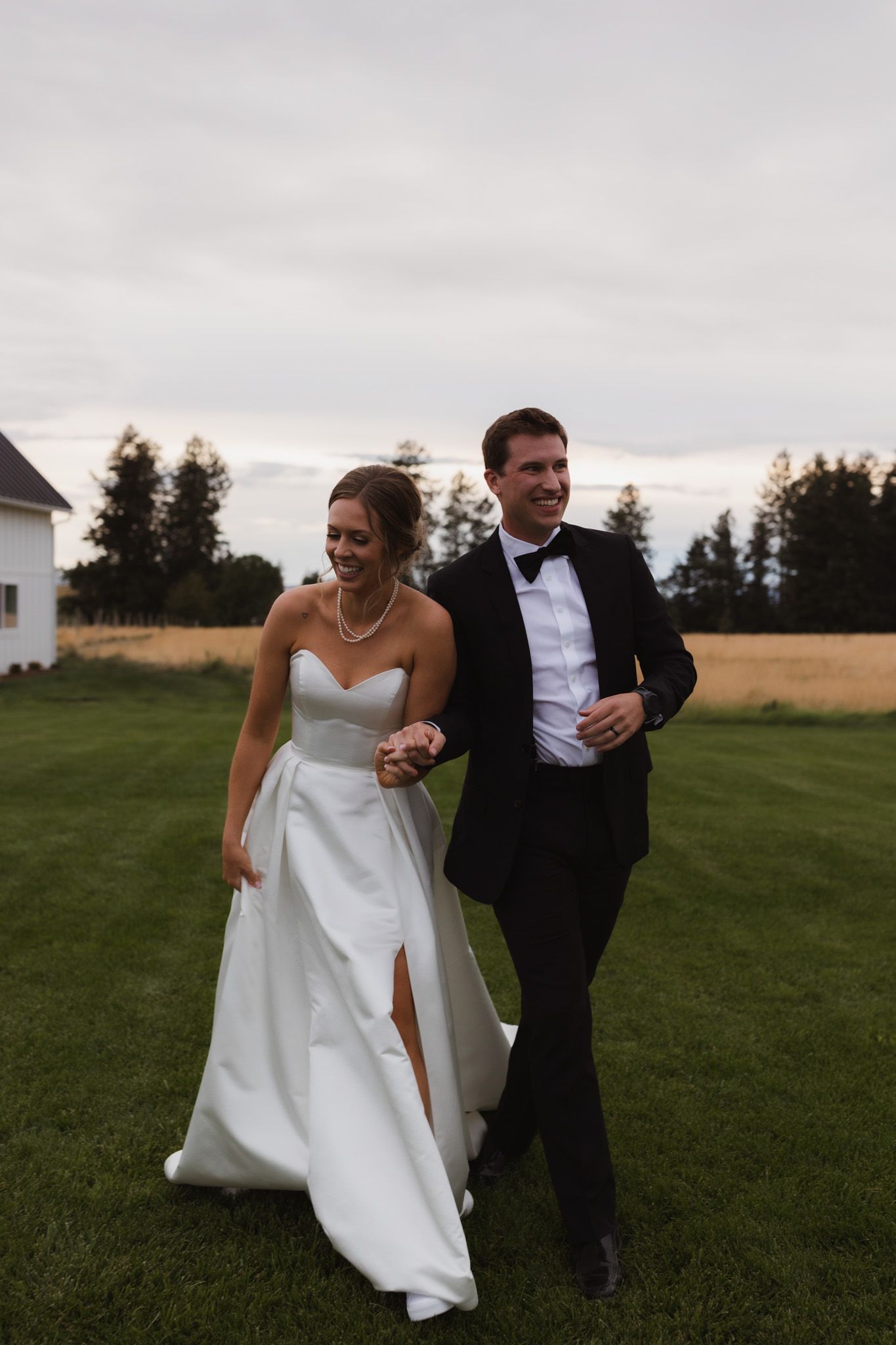 A couple walks together on a grassy lawn at dusk, with the bride wearing a white gown with a slit and groom in a tuxedo. Spokane wedding pho