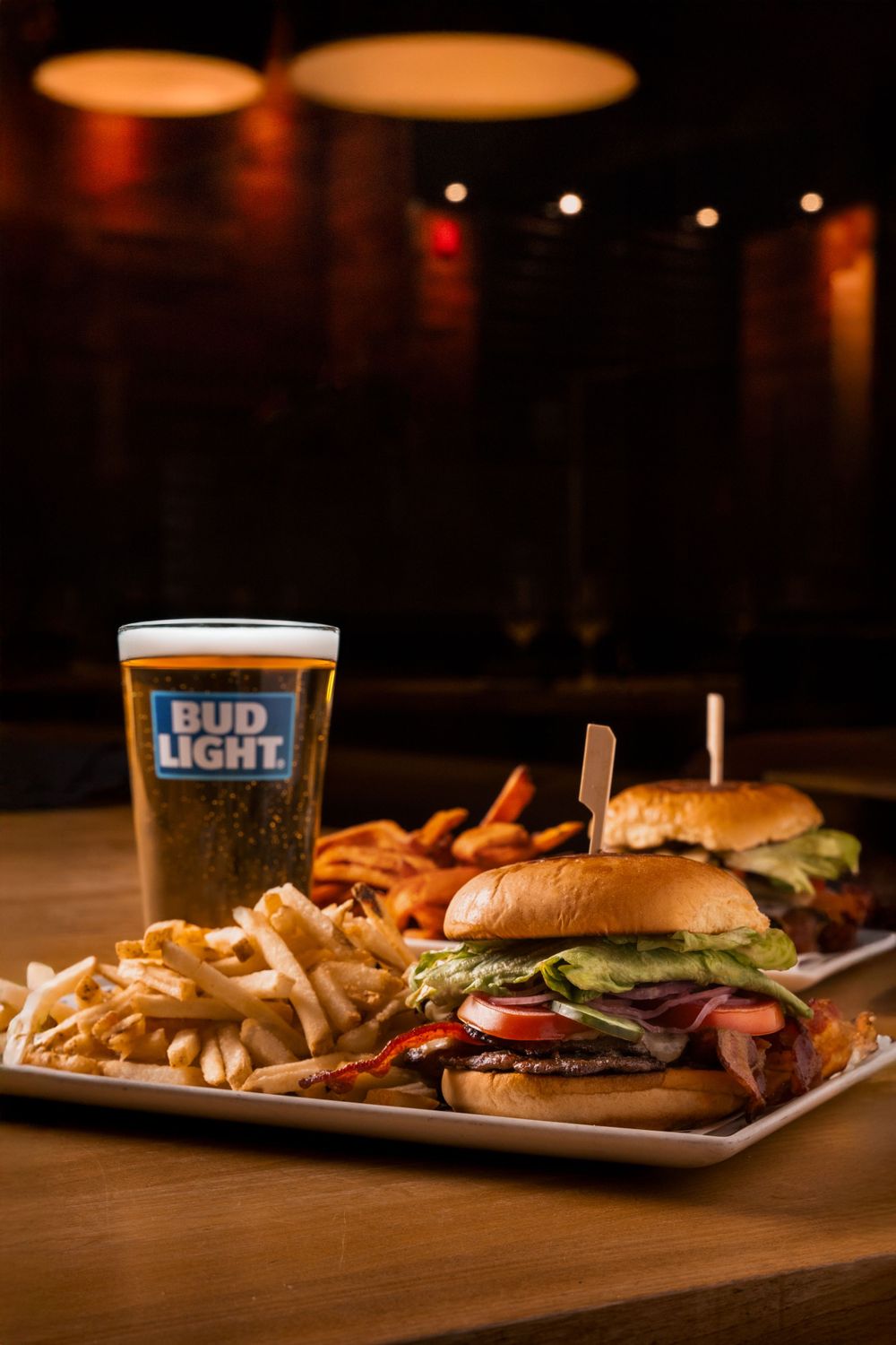 Two juicy hamburgers served with crispy french fries and a cold glass of Bud Light beer on a wooden table in dim lighting.