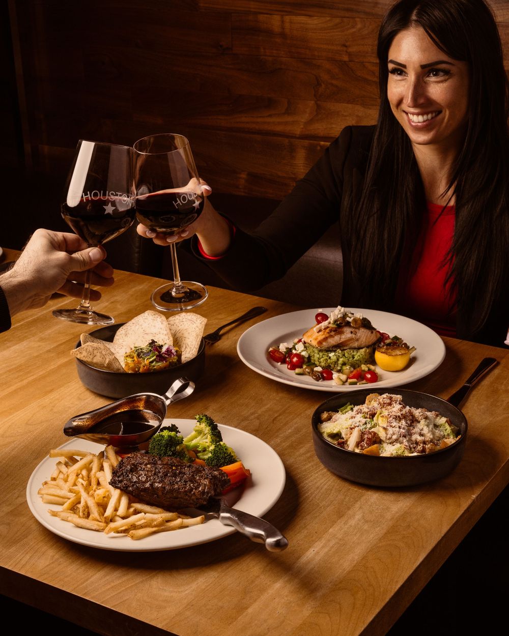 People toasting with red wine glasses over a dinner table spread with steak, fries, and gourmet side dishes at a restaurant.