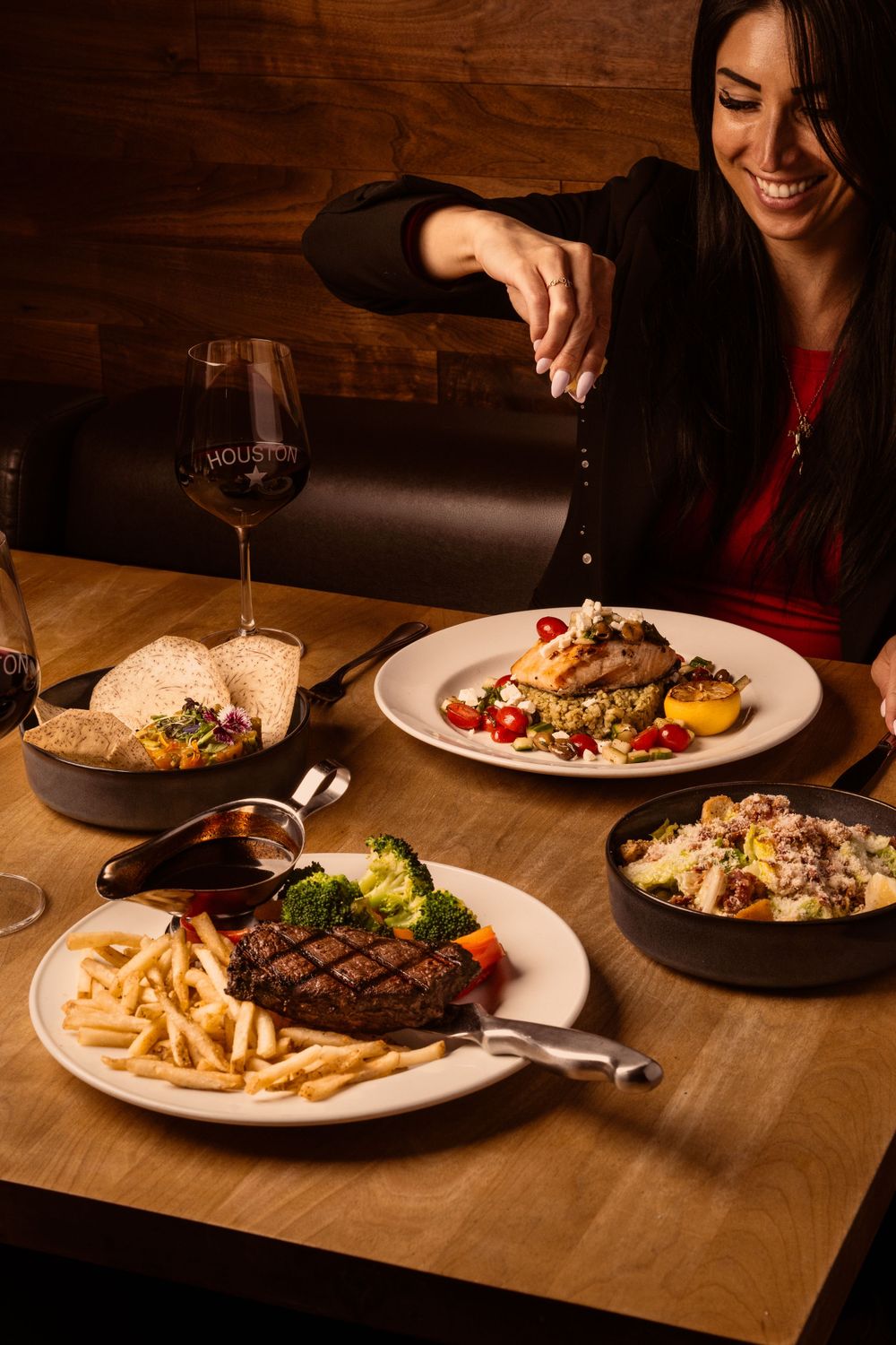 A spread of fine dining dishes including steak with fries, wine, and pasta on a wooden restaurant table in warm lighting.