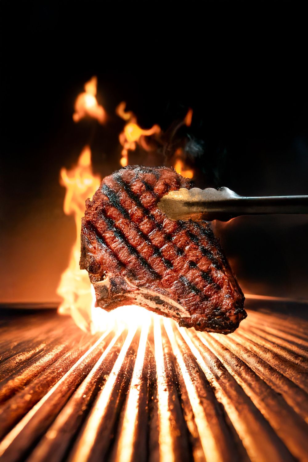 A juicy grilled steak with char marks is held by tongs over flames and grill grates in dramatic lighting.