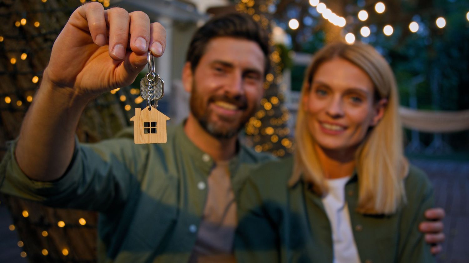 A couple holds up house keys in front of Christmas lights in an evening setting.