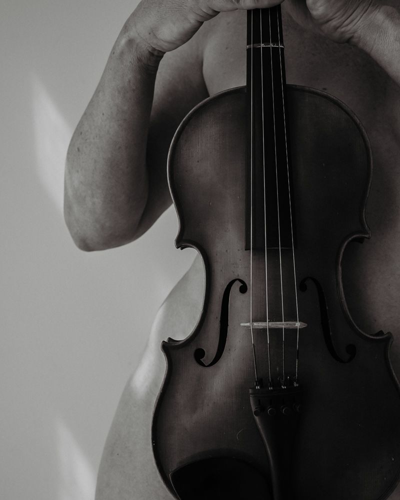 Close-up of violin held against nude torso in dramatic lighting.