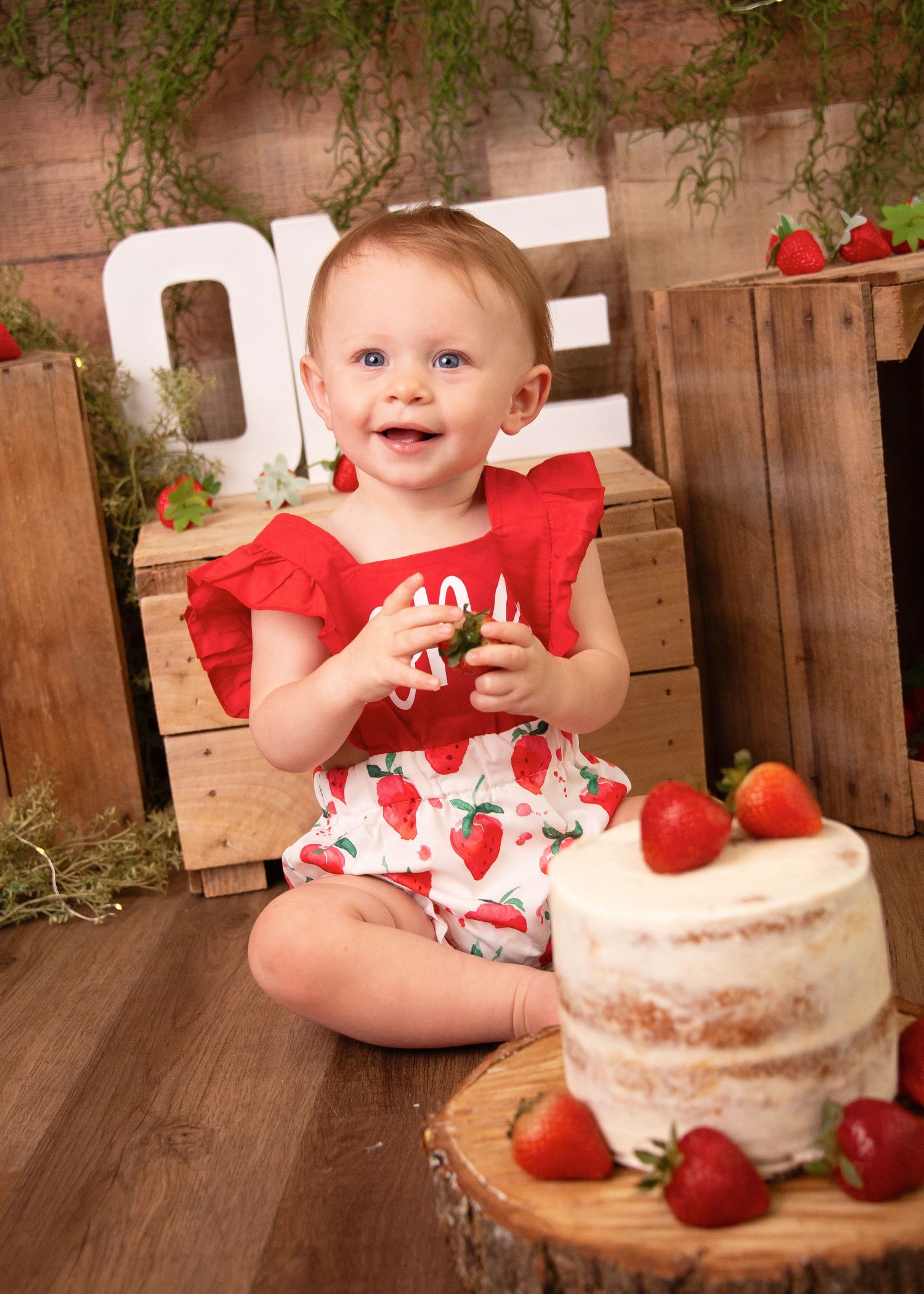 Smiling toddler in red dress next to rustic strawberry cake setup.