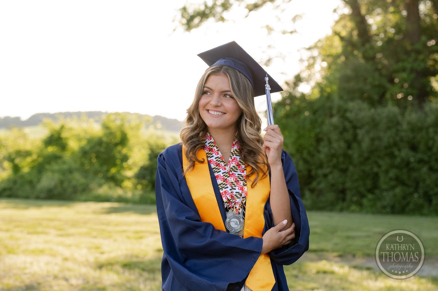 Graduate in traditional academic dress smiles while posing outdoors at sunset.