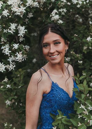 Smiling brunette woman in blue lace prom dress poses among white flowering bushes outdoors. Senior grad photos. Graduation photos.