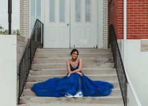 Young woman in royal blue ball gown sits on stone steps outside brick building with white doors. Senior grad photos. Graduation photos.