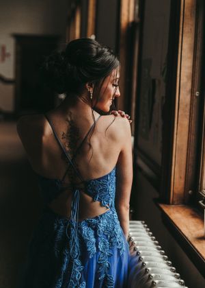 Woman in backless blue lace gown with tattoo gazes out window in moody dramatic portrait. Senior grad photos. Graduation photos.