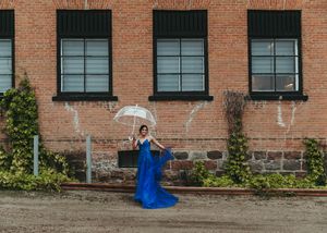 Woman in royal blue gown holds white umbrella outside rustic brick building in outdoor portrait. Senior grad photos. Graduation photos.