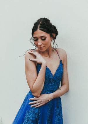 Brunette woman in blue lace gown looks down gracefully against white wall in elegant portrait. Senior grad photos. Graduation photos.