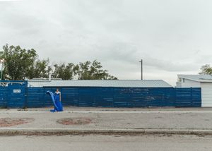 Woman in royal blue formal gown stands before blue industrial fence on urban street under cloudy sky. Senior grad photos. Graduation photos.