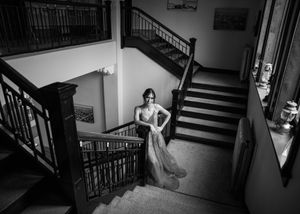 Elegant woman in formal gown poses on grand wooden staircase in black and white portrait. Senior grad photos. Graduation photos.