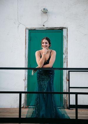 Woman in teal sequin gown smiling on a railing in front of a green door. Senior grad photos. Graduation photos.