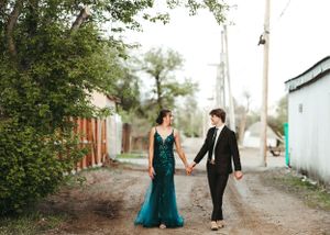 Couple holding hands walking down a tree-lined dirt path, woman in green sequin gown. Senior grad photos. Graduation photos.