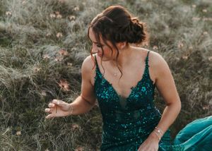 Young woman in emerald green sequin gown sitting in a grassy field, smiling downward. Senior grad photos. Graduation photos.